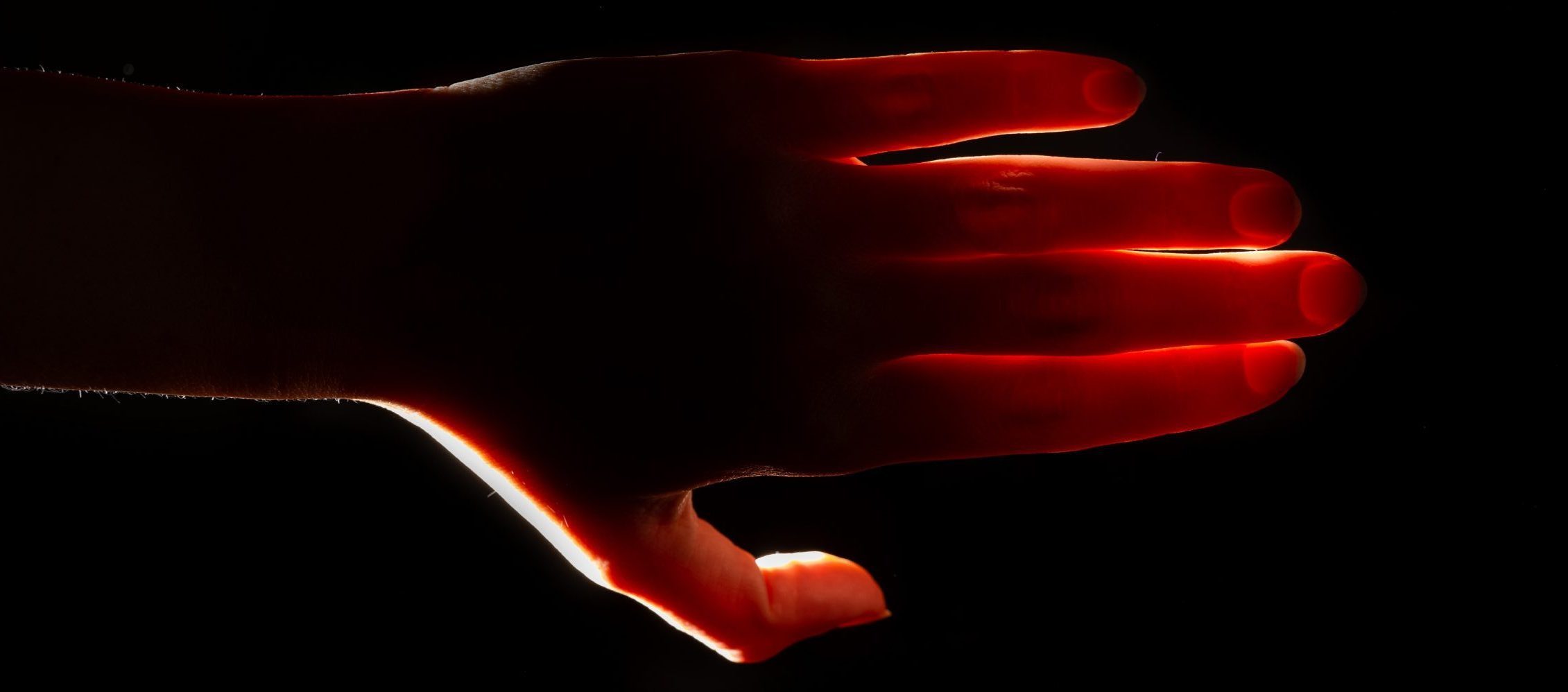 A left hand shown with its fingers outstretched. The hand is in front of a black background, but there is a bright light illuminating the hand from behind.