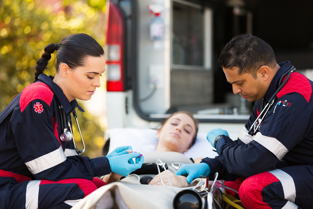 Two EMTs taking a patient's vitals. The patient is lying on a gurney. The EMT workers are wearing red and black uniforms. The open ambulance is in the background.