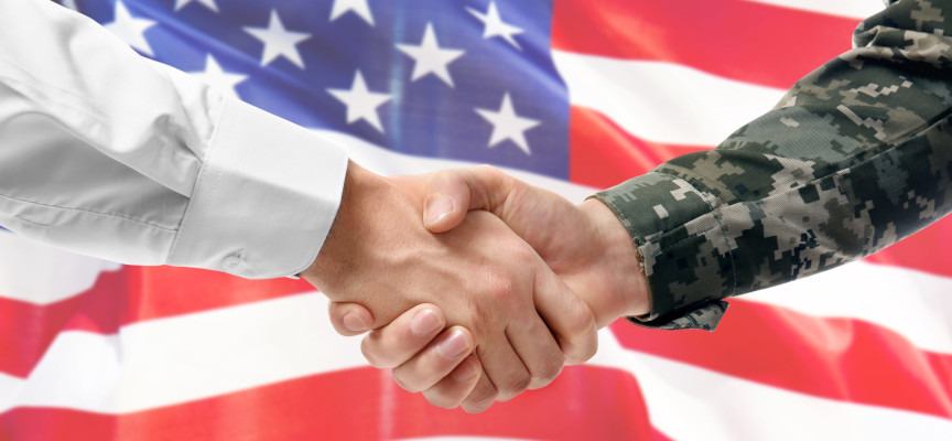 A close up of two hands shaking in front of an American flag. One arm is clothed in a white dress shirt. The other arm is clothed in a military uniform.